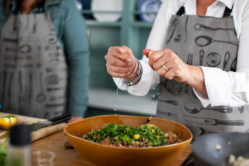 Person preparing a salad with another person in the background, wearing aprons.